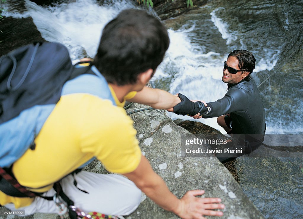 Young Man Pulling up Another Young Man Wearing a Wet Suit From a Waterfall