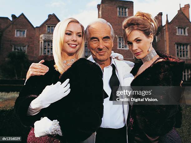 portrait of a wealthy senior man standing between two young women - manteau de fourrure photos et images de collection