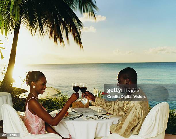 romantic couple sitting in a restaurant on the coast at sunset toasting with glasses of red wine - black couple dining stockfoto's en -beelden
