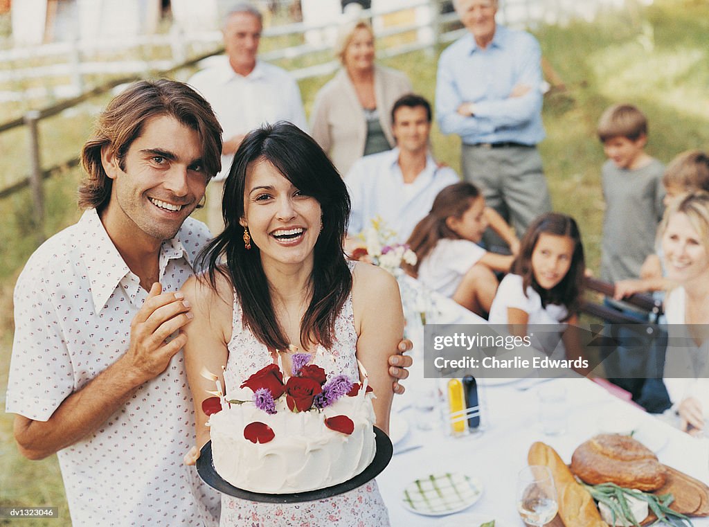 Portrait of a Man Outdoors With His Arm Around a Woman Holding a Birthday Cake and Family in the Background Sitting at a Table
