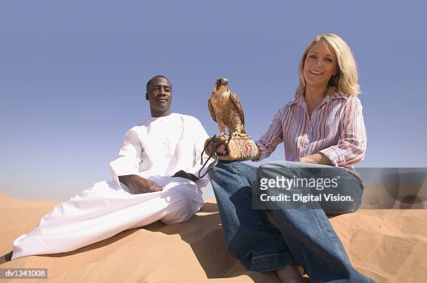 portrait of a woman with a falcon perching on her arm sitting on a sand dune side by side with a man in traditional middle eastern dress - falcon fotografías e imágenes de stock