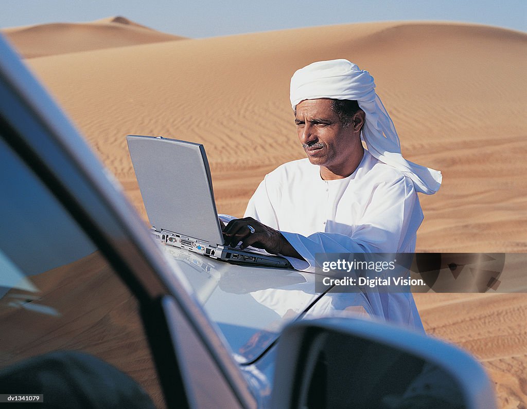Man in Traditional Middle Eastern Dress Using a Laptop on a Car Bonnet in the Desert