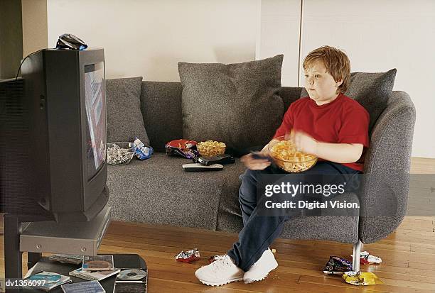 young boy watching tv on a sofa and eating crisps from a bowl - child eat side stock-fotos und bilder