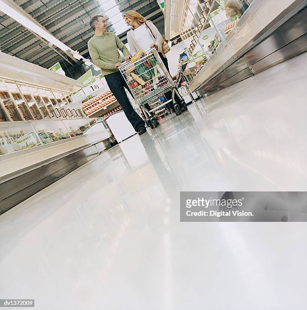 couple walking down a supermarket aisle pushing a full shopping trolley - supermarket trolley female stock-fotos und bilder