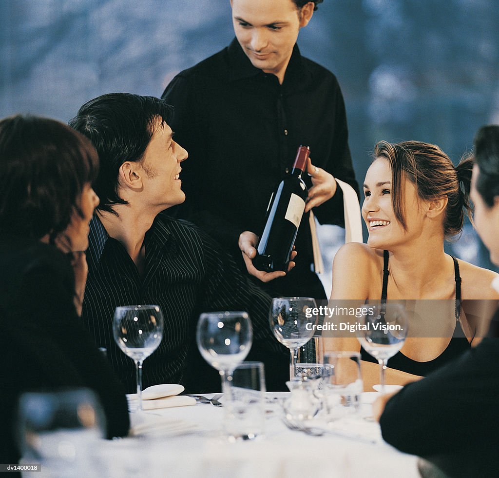 Waiter Showing a Bottle of Red Wine to a Group of Young, Smiling Adults Sitting at a Table at a Restaurant