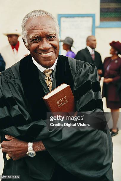 portrait of a smiling priest holding a bible in his crossed arms with people in the background - godsdienstvrijheid stockfoto's en -beelden