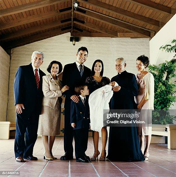 portrait of a three generational family standing in a church with a priest and a baby in a christening gown - christening gown stock pictures, royalty-free photos & images