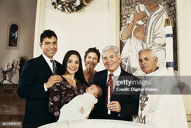 portrait of a family standing by a baptismal font with their baby and a priest - pila bautismal fotografías e imágenes de stock