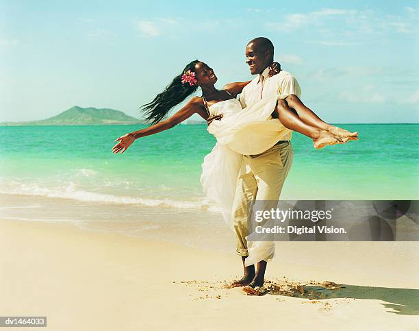 young man carrying his new bride in the sand at the waters edge - black veil brides stock pictures, royalty-free photos & images