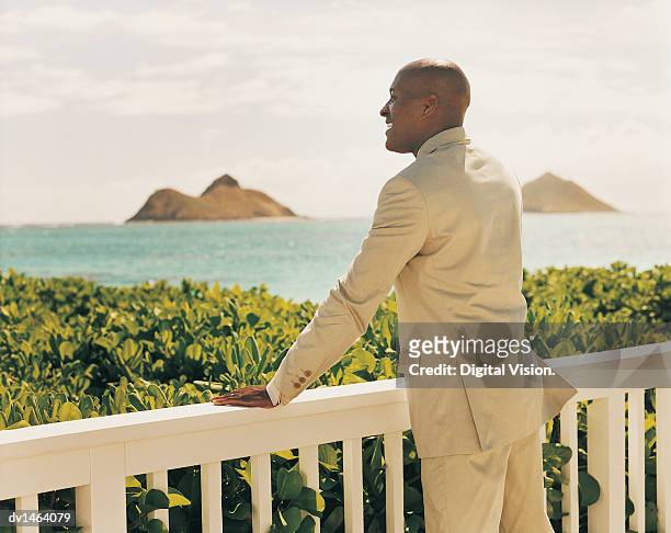 man in a full suit stands leaning on a balcony looking at a tropical view - full suit ストックフォトと画像
