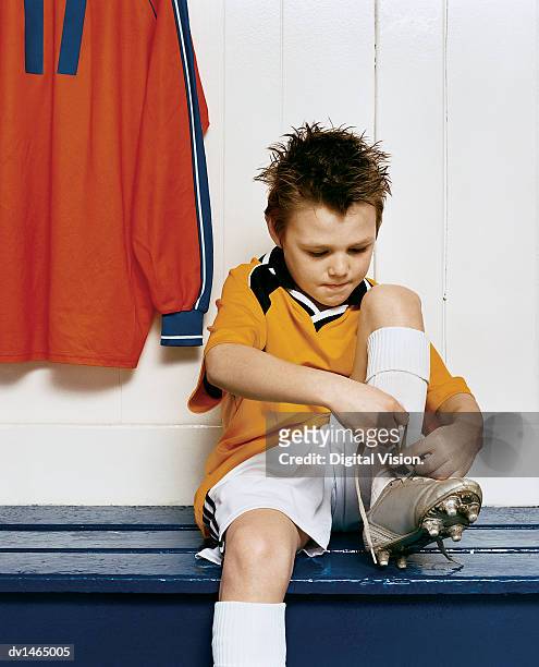 young boy ties the laces of his football boot while sitting on a bench in a changing room - boy tying shoes stock pictures, royalty-free photos & images