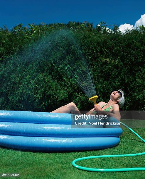 mature woman lies in a paddling pool in a garden, holding a water hose and spraying water - plastic pool stockfoto's en -beelden