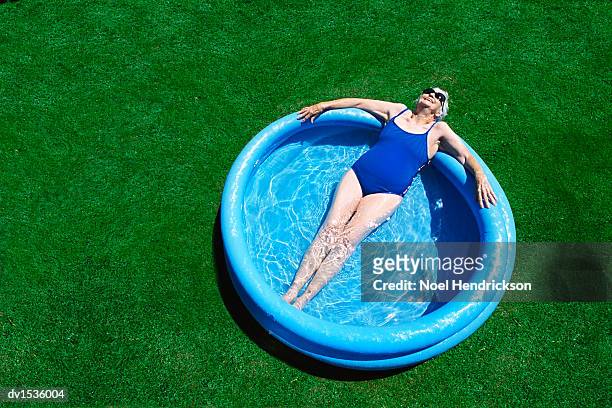 elderly woman lies in a blue paddling pool, sunbathing and relaxing - women sunbathing pool - fotografias e filmes do acervo