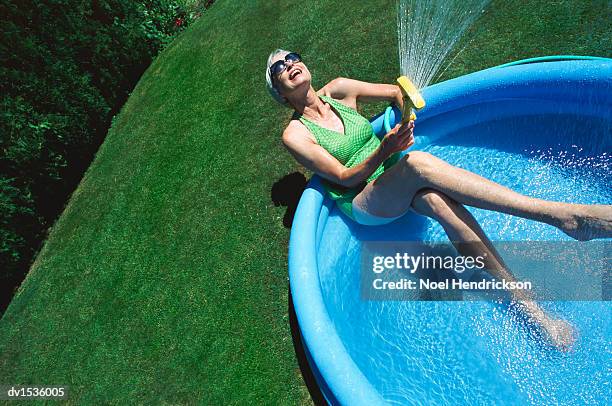 mature woman sits in a paddling pool spraying water from a hosepipe, unusual angle - plastic pool stockfoto's en -beelden