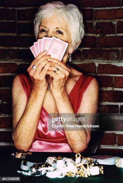 senior woman sitting at a table gambling, holding up playing cards in front of her face - gambling table stock pictures, royalty-free photos & images