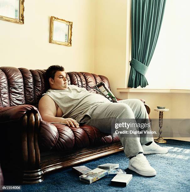 young man lies slouched on a sofa watching videos and holding a packet of crisps - mala postura fotografías e imágenes de stock