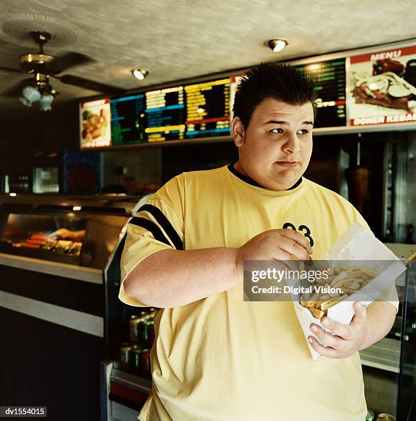 overweight young man stands in a fast food restaurant eating chips from a bag - food bag stock pictures, royalty-free photos & images