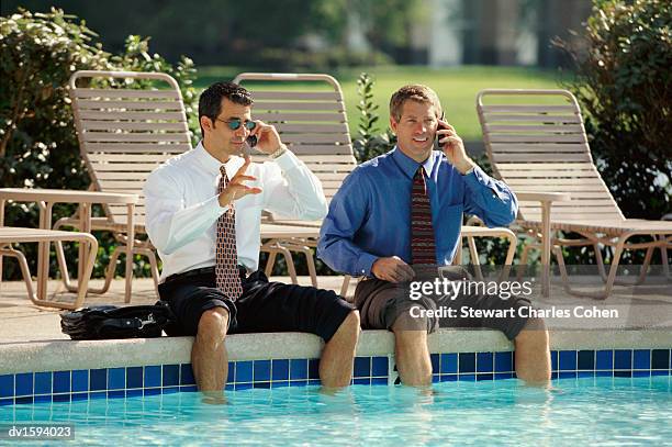 two businessmen with rolled up trousers, sat poolside with their legs in the water, using mobile phones - stewart stock-fotos und bilder