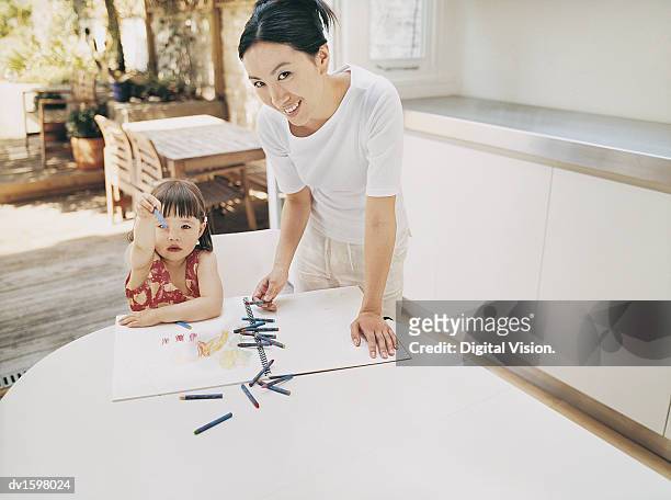 mother standing at a kitchen table by her daughter with her colouring in book and crayons - colouring fotografías e imágenes de stock