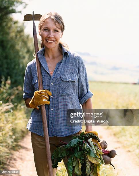 woman standing on a rural dirt track holding a hoe and vegetables - bäuerin stock-fotos und bilder