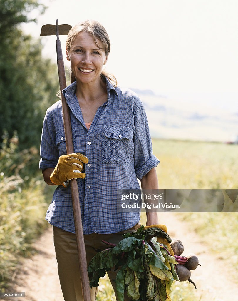 Woman Standing on a Rural Dirt Track Holding a Hoe and Vegetables