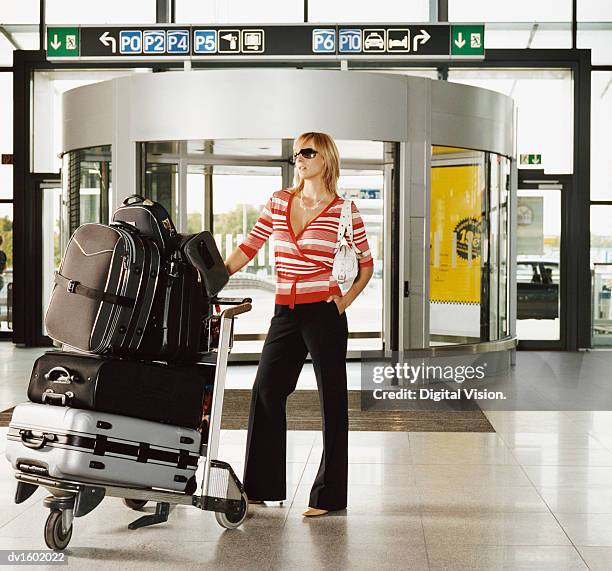 a blond woman standing in an airport with a baggage trolley - carrito-para-equipaje fotografías e imágenes de stock