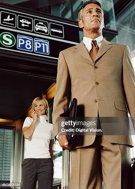 man standing in an airport, wearing a full suit, with a woman standing behind, using a mobile phone - full suit ストックフォトと画像