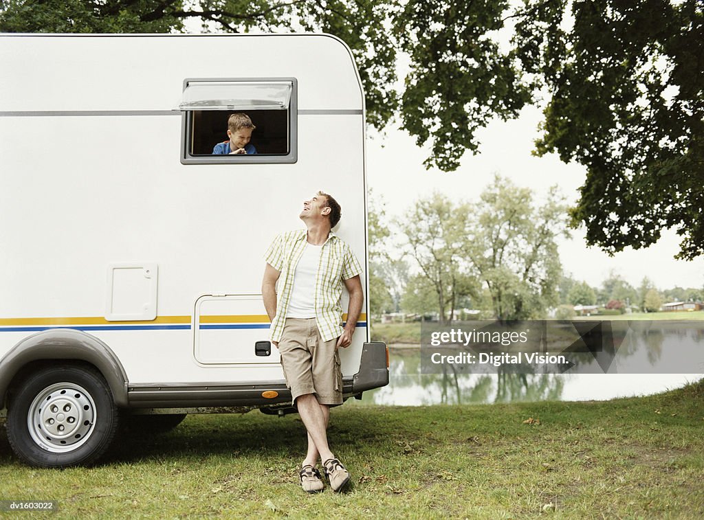 Man Leaning Against His Motor Home Parked Next to a Lake, Looking up at His Young Son Leaning out of the Window