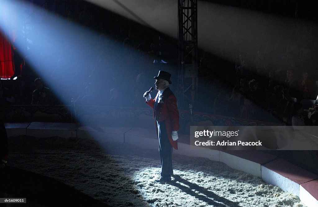 Male Ringmaster Stands in a Spot lit Circus Ring, Making an Announcement to the Audience