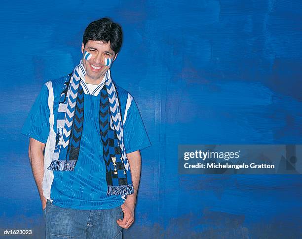 portrait of a smiling, male football supporter standing by a blue wall with his hand in his pocket - supporter scarf stock pictures, royalty-free photos & images