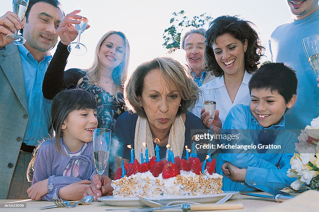 Senior Woman Outdoors Blowing Out Candles on Her Birthday Cake Surrounded by Her Family