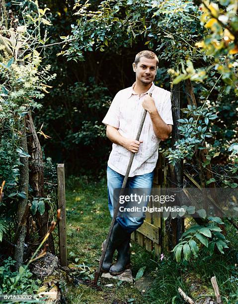 man holding a rake, leaning against a gate post in a rustic garden - rake stock-fotos und bilder