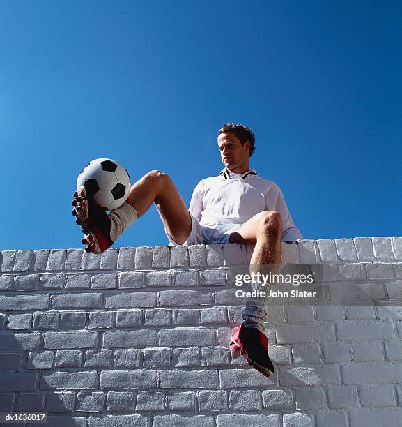 football player sits on a white brick wall holding up a football - football player imagens e fotografias de stock