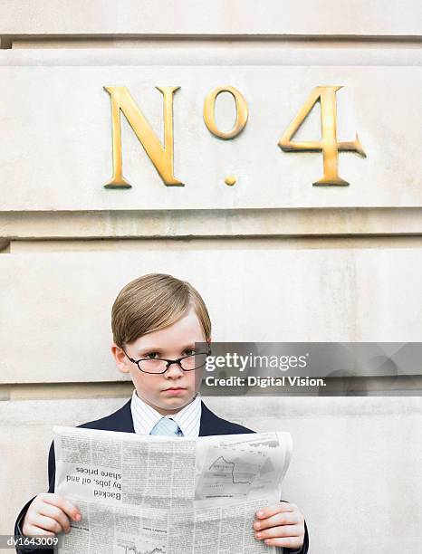 young boy dressed as a businessman reads a financial newspaper below a street number sign - nachahmung erwachsener stock-fotos und bilder