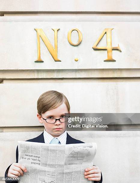 young boy dressed as a businessman reads a financial newspaper below a street number sign - adult imitation stock pictures, royalty-free photos & images