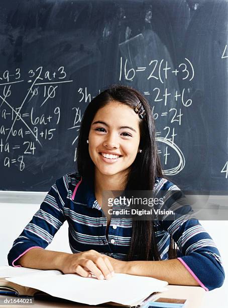 teenage girl sits smiling at a desk in front of a blackboard - girl desk stock pictures, royalty-free photos & images