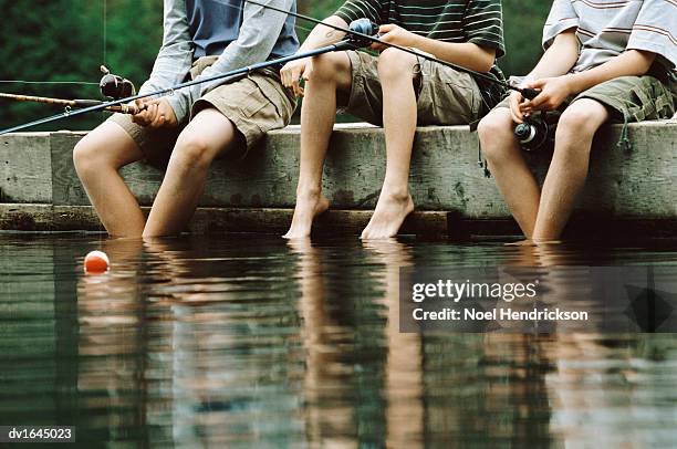 low section of three young boys sitting at the edge of a lake holding fishing rods - angel brinks stock-fotos und bilder