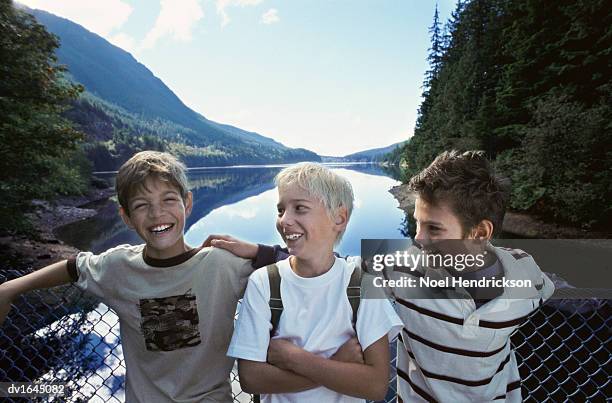 three young boys stand side by side on a footbridge over a lake in a valley, laughing - valley side stock-fotos und bilder