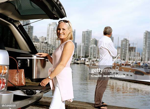 woman unloading a cooler box from an suv vehicle and her partner standing at the water's edge looking at a city skyline view - box purse foto e immagini stock