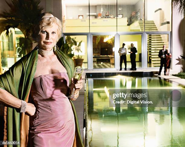senior woman in evening wear holding a glass of champagne stands at the poolside of her mansion home, her dinner party guests in the background - evening wear stock pictures, royalty-free photos & images