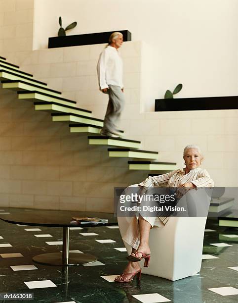 confident well-dressed senior woman sits in an armchair in her modern living room, her husband walking down the stairs - down blouse stockfoto's en -beelden