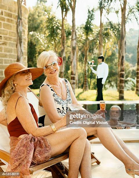 two elegant senior women in swimsuits sit by a pool, a senior man in the pool and their butler in the background - digital butler stockfoto's en -beelden