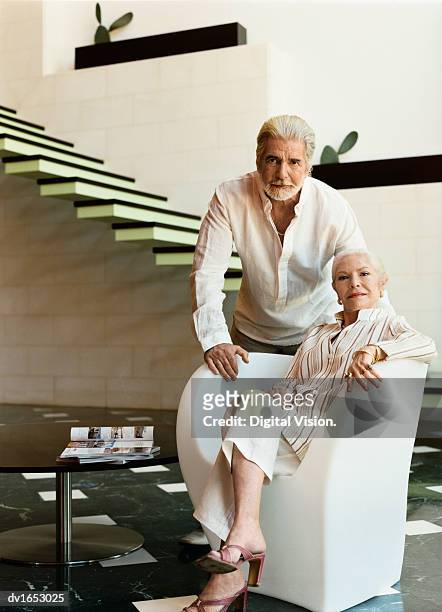 elegant senior woman sitting in an armchair by a coffee table in a modern home, with her husband - elegance is an attitude ストックフォトと画像