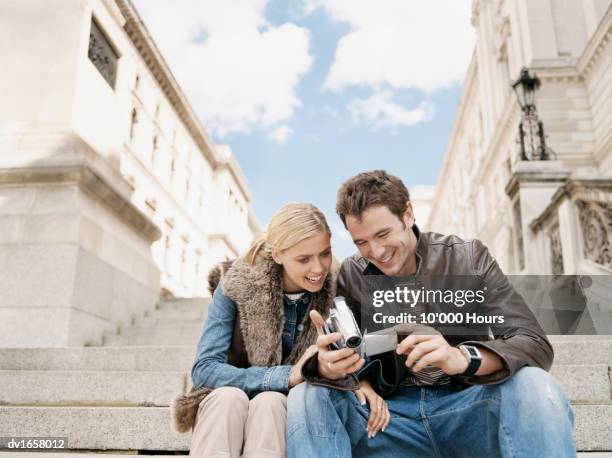 couple sitting side by side on stone steps looking at a camcorder - hours around the world stock pictures, royalty-free photos & images