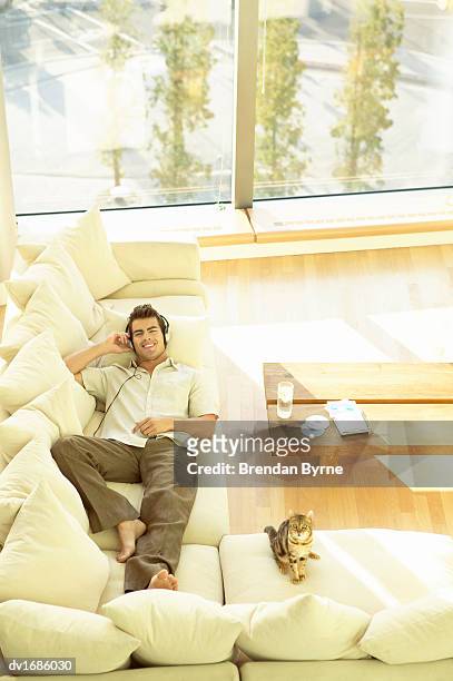 thirty something man lying on a sofa listening to music on headphones with his pet cat - penthouse pet stock pictures, royalty-free photos & images