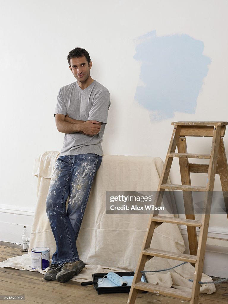 Man Leaning Against a White Wall in a Room Prepared for Decoration, Next to a Step Ladder and Paint Roller