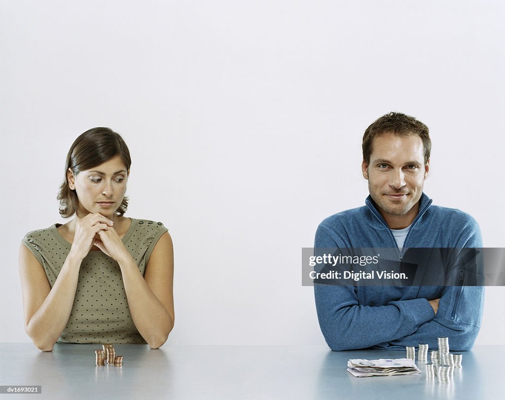 Man and Woman Sit Next to each Other With a Pile of Money in Front of Them, Woman Looking Jealous at Man's Pile