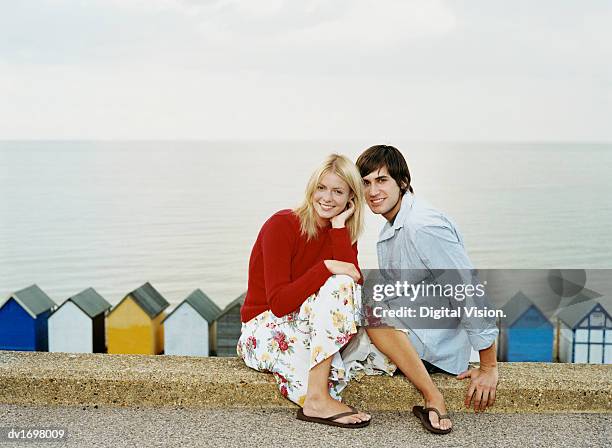 couple sat on a kerb at the seaside with beach huts in the background - kerb stock-fotos und bilder