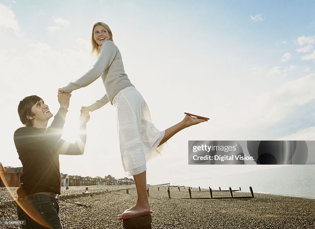 Man Holding Hands with a Woman as she Balances on a Breakwater Post on a Pebble Beach