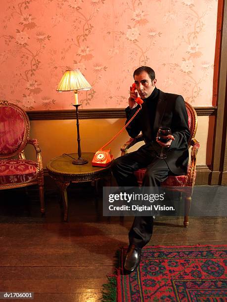 well-dressed man sits in an ornate armchair holding a glass of red wine and making a phone call - ornate house furniture stock-fotos und bilder